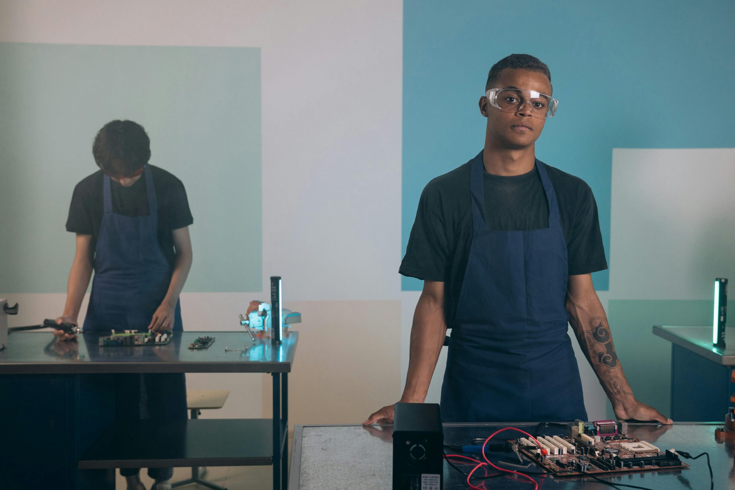 Two men working on electronics in a workshop, wearing aprons and safety goggles.